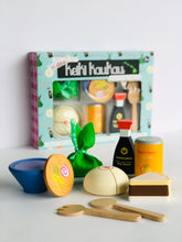 Load image into Gallery viewer, Teal box reading "More! Keiki Kaukua" behind wooden play food set (saimin, laulau, shoyu bottle, passion orange juice can, manapua, chocolate haupia pie, a fork and a spoon)