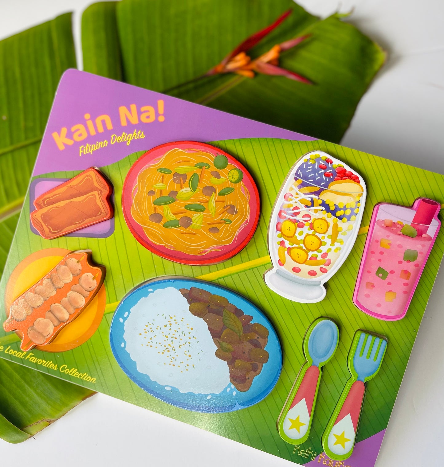 Wooden Puzzle reading "Kain Na! Filipino Delights" with lumpia, pancit, halo-halo, adobo, barbecue, a pink drink, and utensils on a green background.
