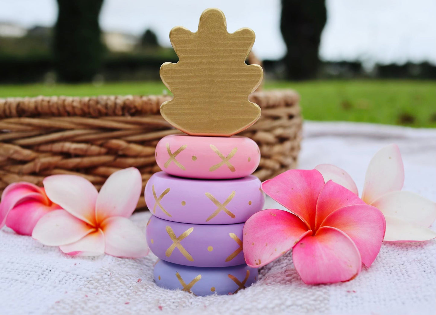 Pink, purple, and gold wooden pineapple stacking toy next to plumeria flowers