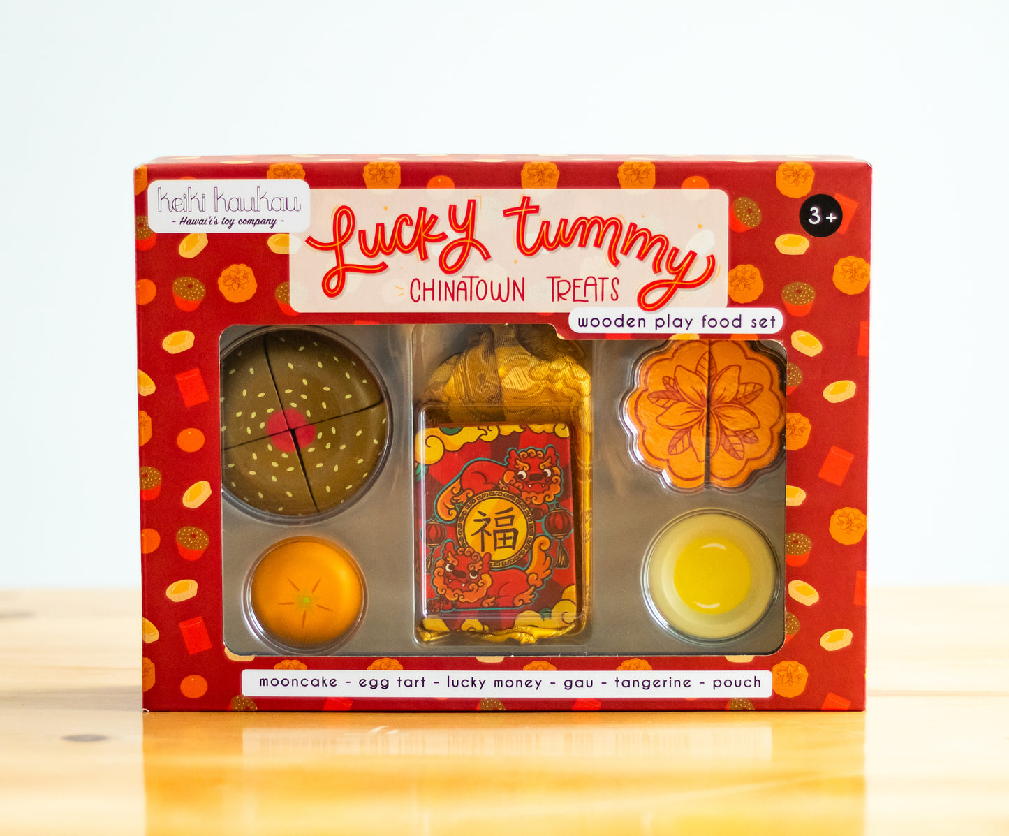 Red box labeled "Lucky tummy Chinatown treats wooden play food set" showing play food contents (mooncake with plumeria design, 4 piece gau, egg tart, tangerine, red lucky envelope, and golden storage pouch)