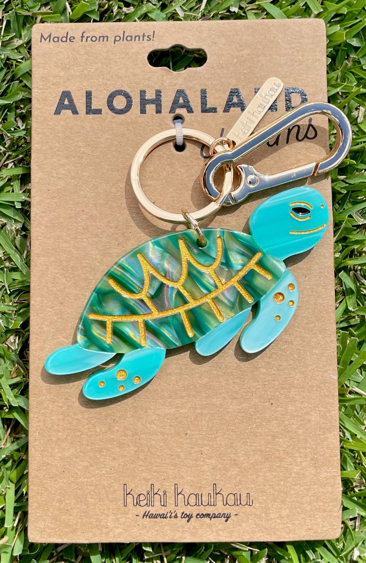 Turtle-shaped keychain on a brown card with 'Aloha Land' branding, displayed on grass.