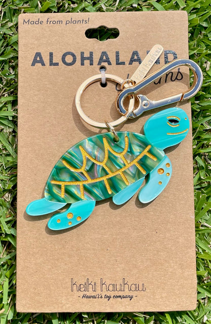 Turtle-shaped keychain on a brown card with 'Aloha Land' branding, displayed on grass.