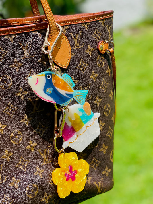 Colorful fish keychain attached to a brown patterned handbag with a blurred green background