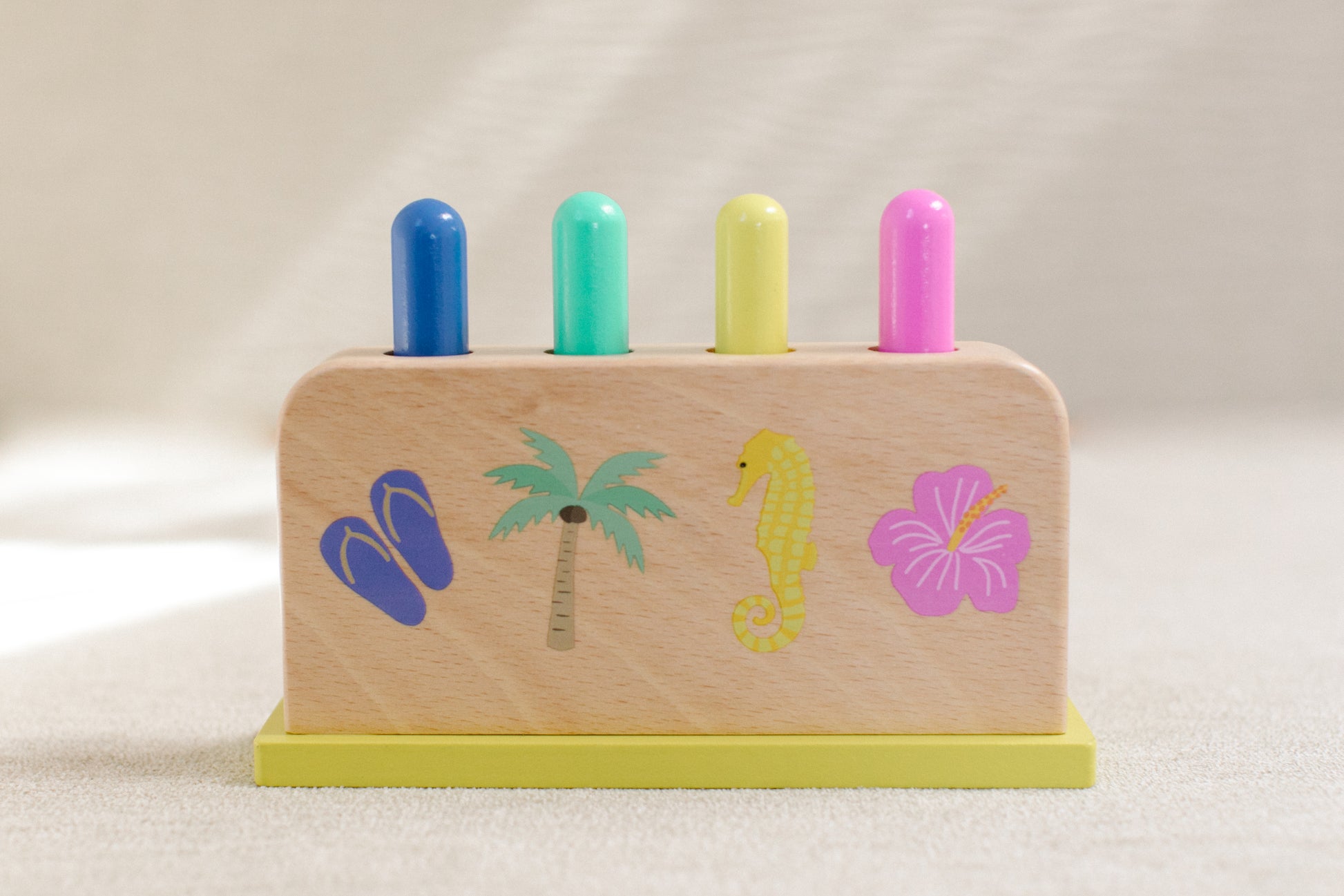 Blue, teal, yellow, and pink pegs inside of wooden jumping pegs box styled with slipper, coconut tree, seahorse, and hibiscus graphics