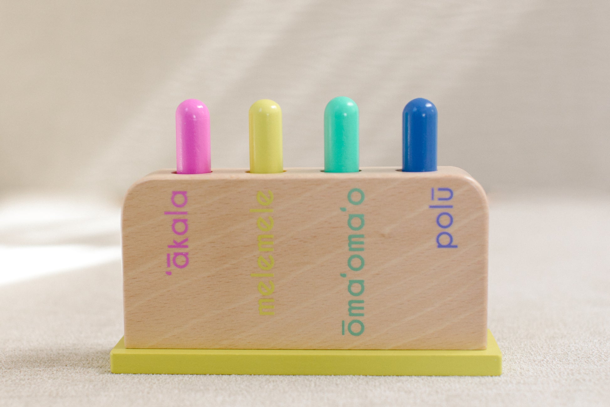 Wooden jumping peg box reading "‘ākala, melemele, ōma‘ōma‘o, polū" with all four pegs