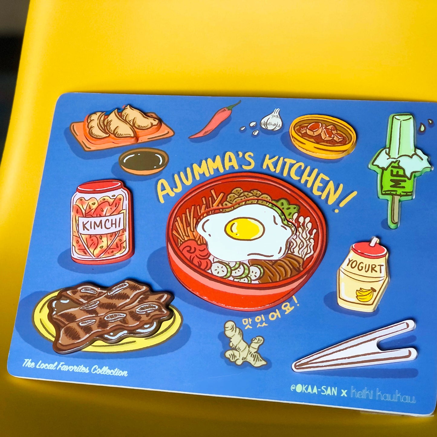 Blue wooden puzzle with 8 block pieces (mandoo, gamja jorim, melon ice cream bar, kimchi jar, bibimbap, banana milk drink, kalbi, and chop sticks) reading "Ajumma's Kitchen!"