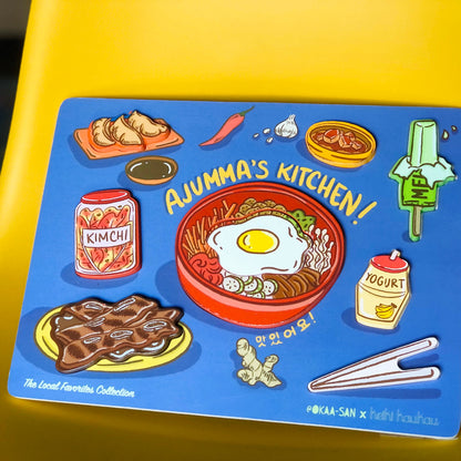Blue wooden puzzle with 8 block pieces (mandoo, gamja jorim, melon ice cream bar, kimchi jar, bibimbap, banana milk drink, kalbi, and chop sticks) reading "Ajumma's Kitchen!"
