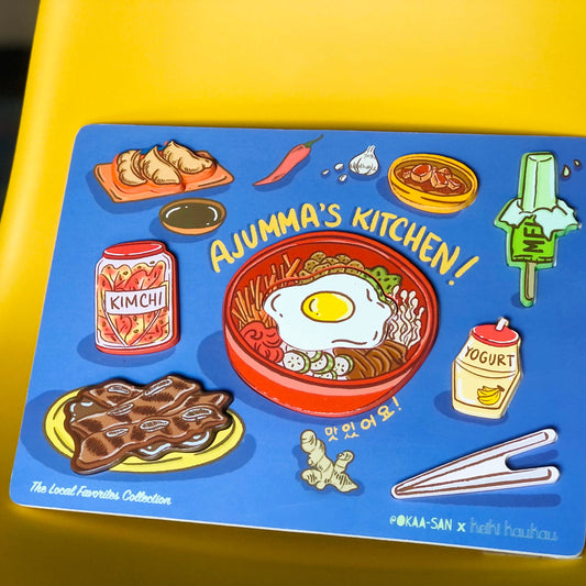 Blue wooden puzzle with 8 block pieces (mandoo, gamja jorim, melon ice cream bar, kimchi jar, bibimbap, banana milk drink, kalbi, and chop sticks) reading "Ajumma's Kitchen!"