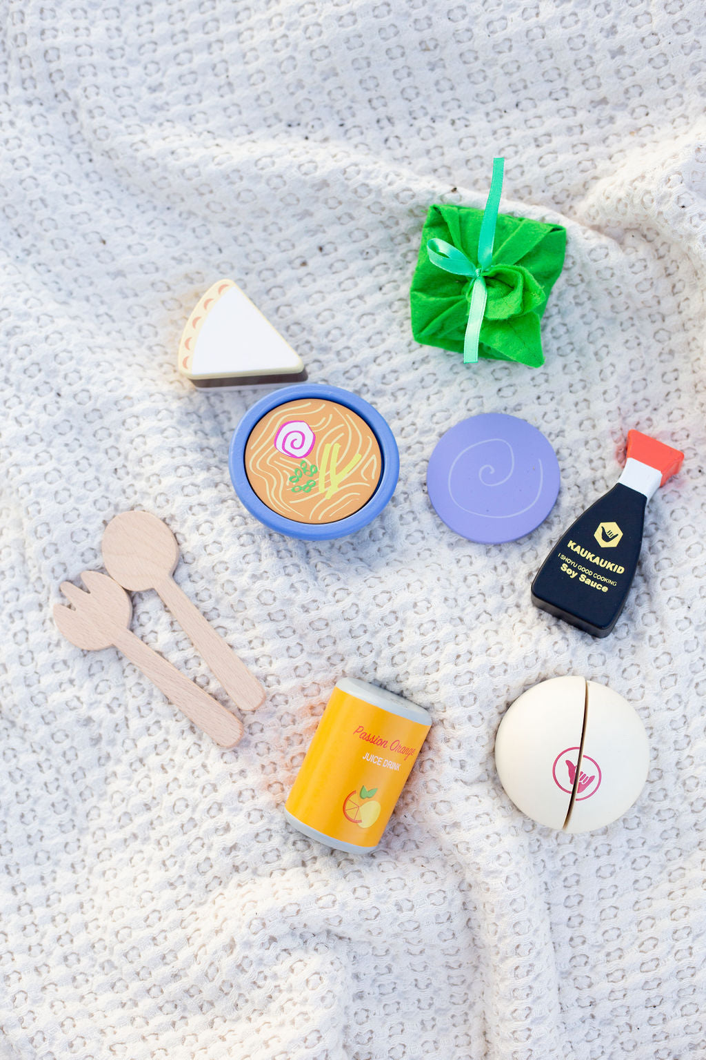 Wooden play food toy set (fork, spoon, chocolate haupia pie, saimin, passion orange juice can, poi, laulau, shoyu bottle, and a manapua) on a white blanket