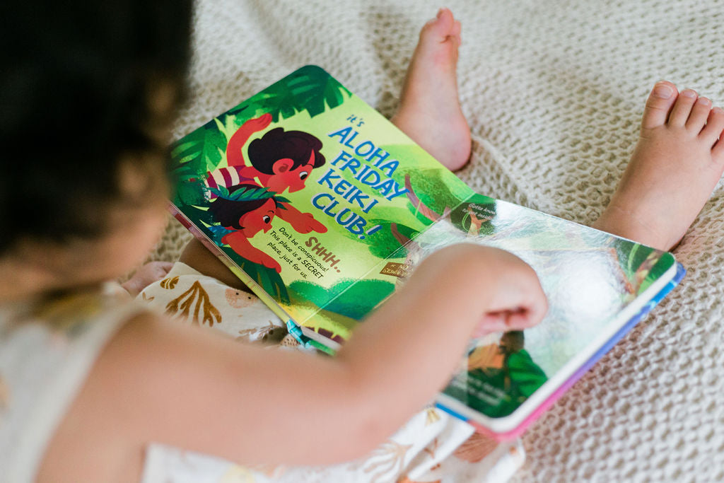 Toddler pointing to an open page of "The Aloha Friday Keiki Club" Book reading "it's Aloha Friday Keiki Club"