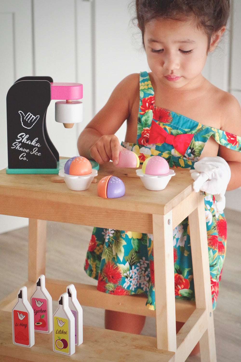 Little girl playing with wooden shave ice pieces next to wooden shave ice machine and syrup bottles