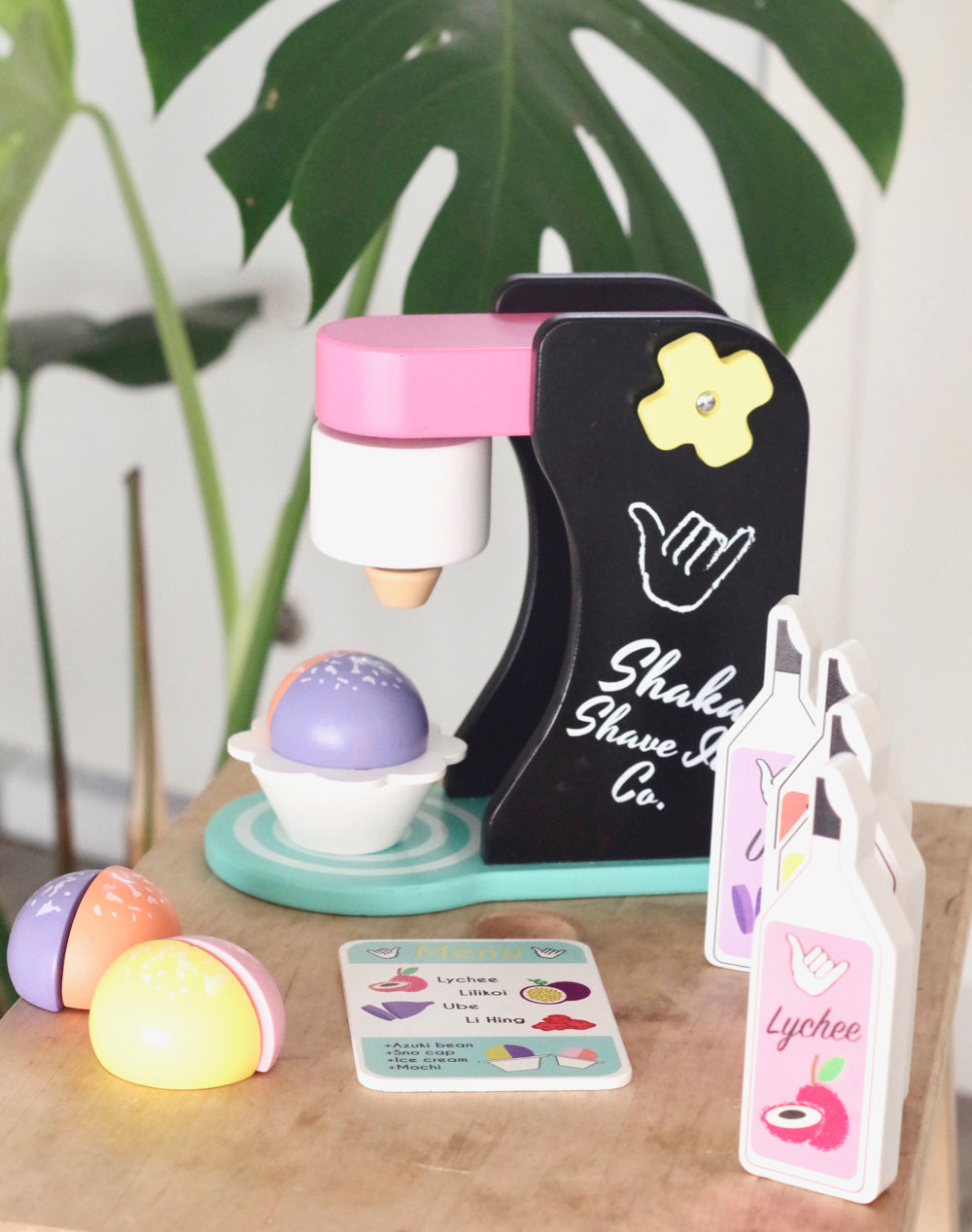 Black and pink wooden toy shave ice machine with toy syrup bottles, menu, and purple, pink, and orange shave ice pieces.