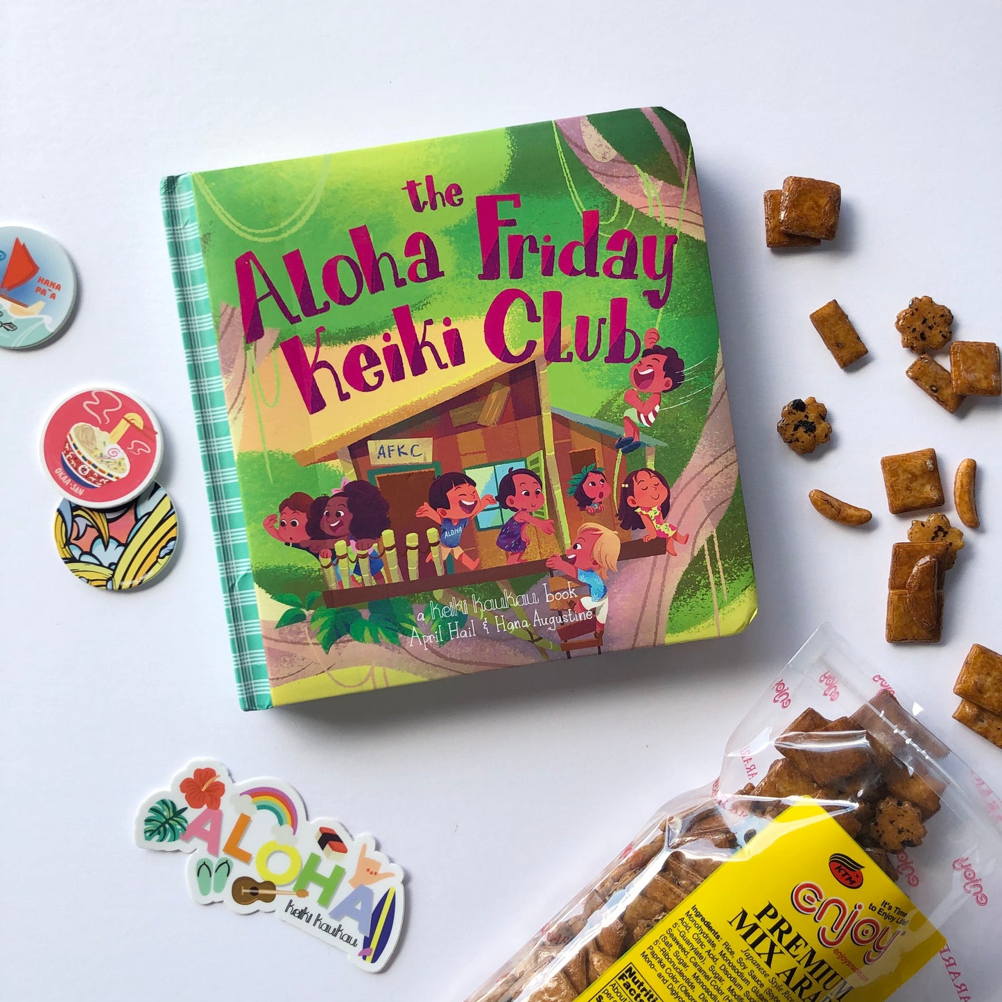 Book cover with illustration of kids in a tree house that reads "The Aloha Friday Keiki Club" next to spilled arare snack, milk caps, and a sticker reading "Aloha"