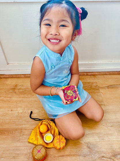 Little girl in blue Chinese dress sitting next to golden pouch and play food while holding red lucky envelope