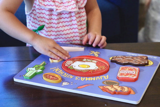 Little girl placing banana milk wooden puzzle piece into blue puzzle reading "Ajumma's Kitchen"