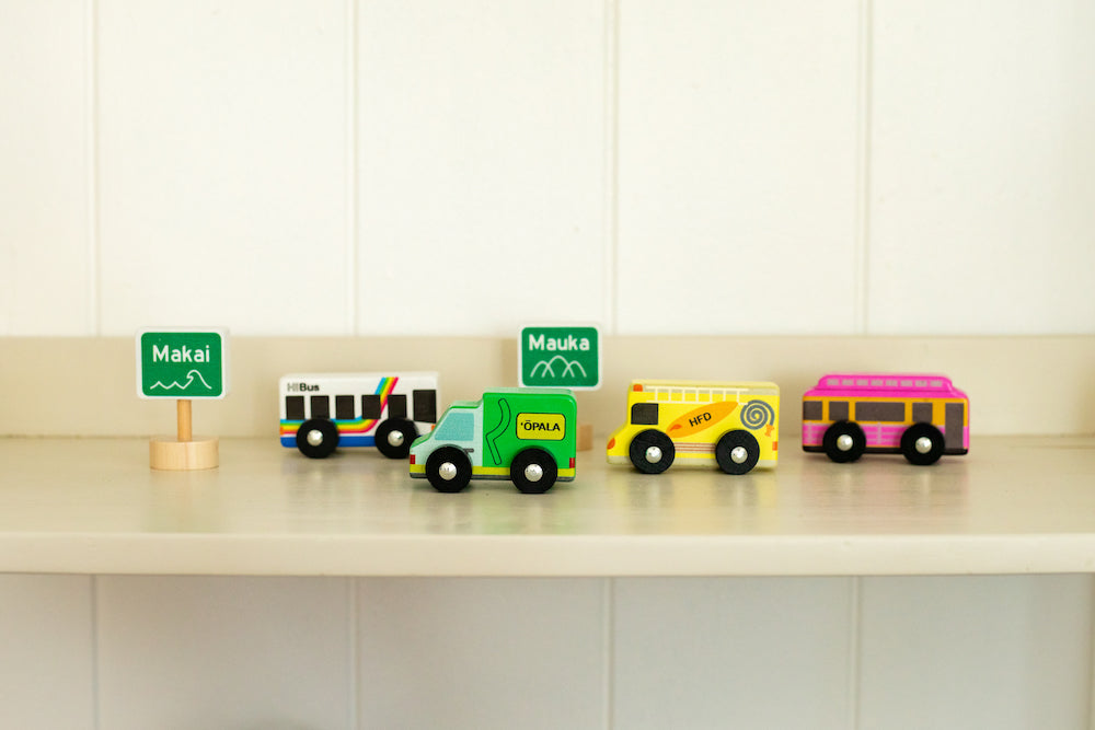 Four wooden cars (rainbow bus, green garbage truck, yellow fire truck, and pink trolly) with two wooden street signs on a shelf