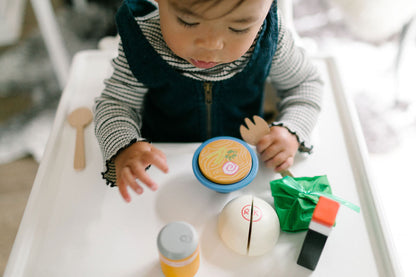 Baby holding wooden fork and playing with wooden play food toys (saimin, passion orange juice can, manapua, laulau, and shoyu bottle)
