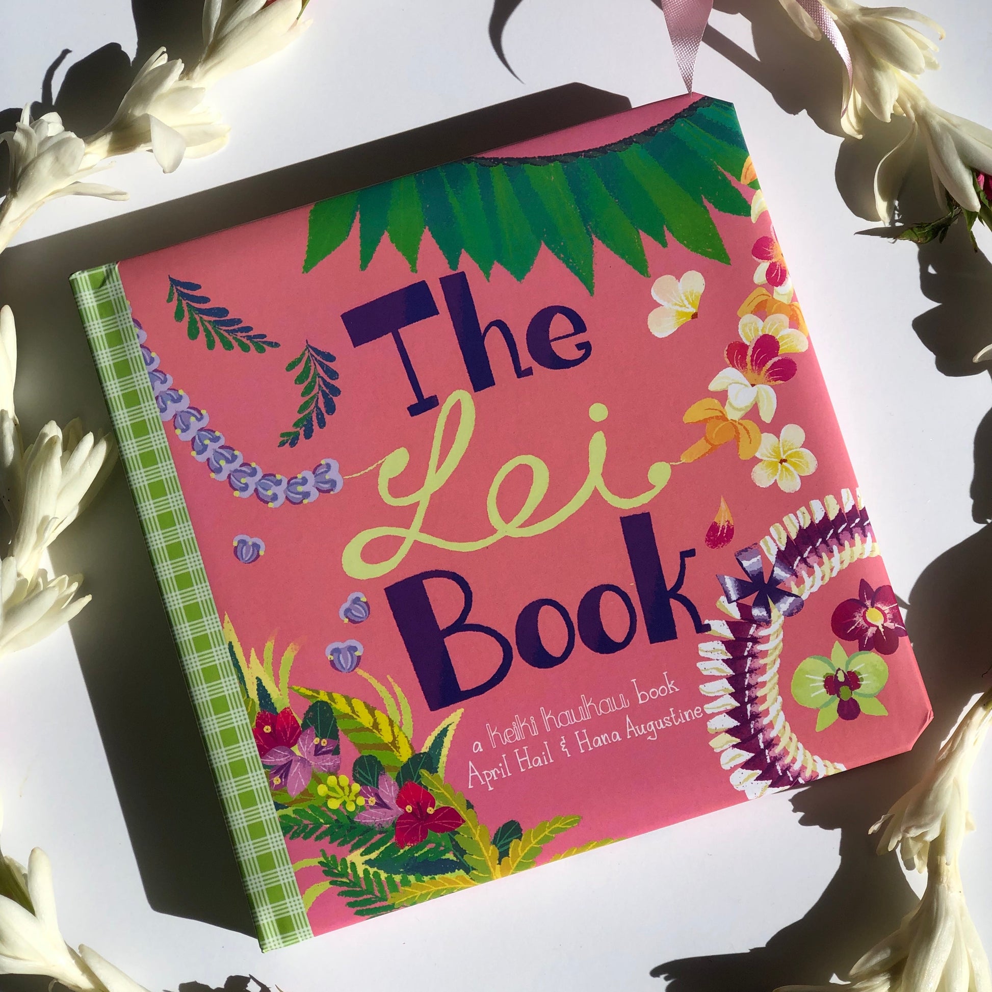 Cover of a pink book with green palaka print spine and illustrations of different flora leis reading "The Lei Book"